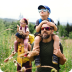 Two young adults carry kids on their shoulders through a field of flowers.